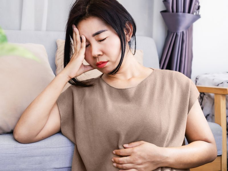 Asian American woman with her hand on her head and her stomach in beige top worry about what to expect from a hysterectomy