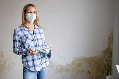 pretty young woman standing in front of wall with mold with tablet and mouth nose mask
