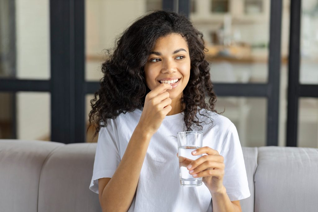 Young beautiful smiling african American woman holding holding vitamin pill and glass of water sitting on the couch at home. Healthy lifestyle, healthy diet nutrition concept
